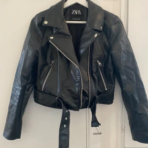 Svart skinnjacka från Zara - Säljer en svart skinnjacka från Zara med bälte i midjan. Den är använd några gånger. Öglan man hänger jackan i är sönder men inget som syns när den sitter på