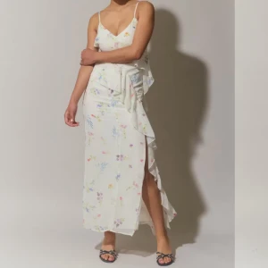 Vit långklänning med blommigt mönster och volang - Säljer en romantisk vit långklänning med smala axelband och blommigt mönster i pastellfärger. Klänningen har volangdetaljer längs framsidan och är tillverkad i ett lätt, mjukt material. Perfekt för sommarens alla tillfällen! Aldrig använt! 