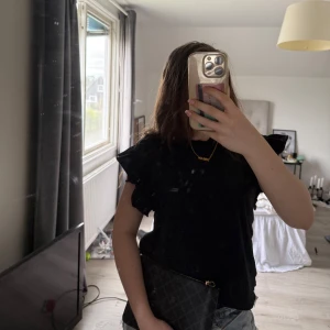 Svart volangtopp - Säljer en svart topp med volangdetaljer på ärmarna. Toppen har en klassisk rund halsringning och är tillverkad i ett mjukt material. Perfekt att styla med jeans eller kjol för en trendig look.