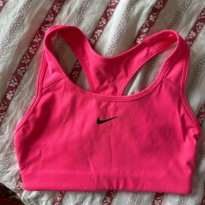 Rosa sport-bh från Nike - Säljer en starkt rosa sport-bh från Nike med klassisk brottarrygg och svart logga framtill. Perfekt för träning och ger bra stöd. Mjukt och stretchigt material som sitter skönt mot kroppen.