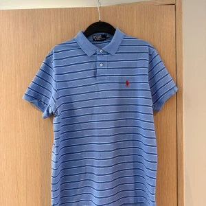 Blå randig pikétröja från Polo Ralph Lauren - Säljer en klassisk blå pikétröja från Polo Ralph Lauren med vita och mörkblå ränder. Där är något smuts på sidan 4 men det går säkerligen bort i tvätten. Nypris ligger på ca 1500