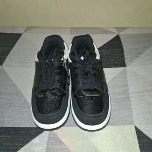 Svarta och vita sneakers från 157 - Säljer ett par svarta och vita sneakers från 157 med klassisk design. Skorna har snörning, rund tå och platt sula. Ovandelen är i syntetmaterial med vita detaljer på sidorna och svart bas. Perfekta för en stilren vardagslook.