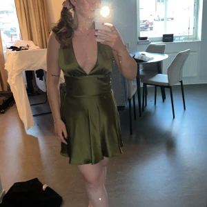 Olivgrön satin klänning med djup v-ringning - Säljer en helt oanvänd olivgrön kortklänning i satin med lappen kvar. Klänningen har djup v-ringning och öppen rygg. Klänningen har knyt i midjan och är ärmlös med en lätt utsvängd kjol. Den är från zara i storlek S