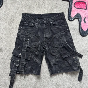 Svarta cargo shorts med detaljer och remmar - Säljer ett par svarta cargo shorts med flera fickor, silverfärgade knappar och dekorativa remmar. Shortsen har råa kanter nertill och coola sömdetaljer över hela plagget. Perfekta för dig som gillar streetwear och unik stil.