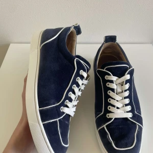 Christian Louboutin skor  - Säljer ett par marinblåa sneakers från Christian Louboutin med vit snörning och vita kanter. Skorna har en klassisk rund tå och platt sula med den ikoniska röda undersidan. Materialet är sammet och de har en sportig men stilren look.