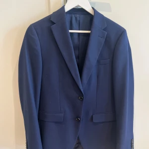 Mörkblå kostym från Riley Stanton - Säljer en stilren mörkblå kostym från Riley Stanton. Kavajen har två knappar, klassiska slag och fickor med lock. Byxorna är raka och matchar kavajen perfekt. Perfekt för bal eller fest. Endast använd 1 gång på student💗🥰