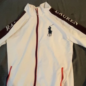 Vit jacka från Ralph Lauren med röd detalj - Säljer en vit jacka från Ralph Lauren med röd dragkedja och röda detaljer vid fickorna. Jackan har lång ärm och logga på bröstet samt text på axlarna. Perfekt för en sportig och stilren look.