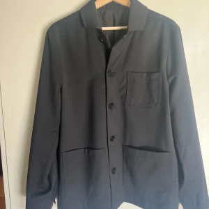 FILIPPA K Louis Wool Gabardine kavajovershirt - Stilren svart kavajovershirt från Filippa k med klassisk krage och tre framfickor. Jackan har knäppning framtill och är perfekt för vår, sommar och höst. Enkel och tidlös design som passar till många olika outfits. Materialet är tunnt och påminner om en kavaj. Nypris: 3399, Säljer för 1500 (Oanvänd)