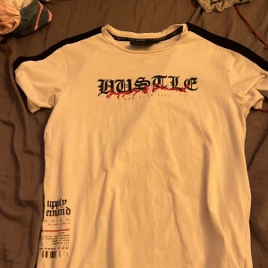 Vit t-shirt med svart text från Supply & Demand - Vit t-shirt från Supply & Demand med svart text 'HUSTLE' på bröstet och tryck nere till vänster. Svarta detaljer längs axlarna. Skön passform och perfekt för en streetwear-look.