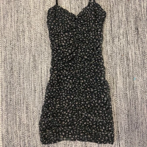 Svart blommig kortklänning - Säljer en svart kortklänning med små blommor på från H&M. Klänningen har smala axelband, åtsittande passform och rynkade detaljer både fram och bak. Den har dubbla tyger som är tunna och luftiga och den glider inte upp 💕