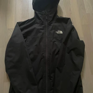 Svart vindjacka från The North Face - Säljer en svart vindjacka från The North Face med huva och dragkedja framtill. Jackan har en enkel och stilren design med logga på bröstet. Perfekt för blåsiga dagar och lätt att packa med sig.