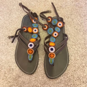 Bruna sandaler med färgglada pärldetaljer - Unika bruna sandaler med färgglada pärlor i blått, orange, vitt och grönt. Remmarna är i skinn och sandalerna har öppen tå och platt sula. Perfekta för varma dagar och ger en bohemisk touch till din outfit.