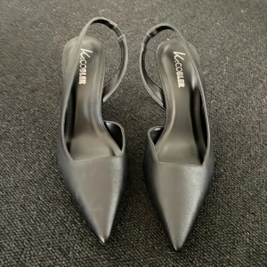Svarta pumps med spetsig tå från LuCober - Säljer ett par eleganta svarta pumps från LuCober med spetsig tå och öppen häl. Skorna har en smal klack och är tillverkade i syntetmaterial med slät finish. Perfekta för dig som vill ha en stilren och klassisk look.