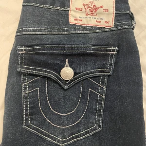 True Religion Becca Bootcut Jeans ni - Mörkblå jeans från True Religion, modell Becca med bootcut och snygga kontrastsömmar. Klassisk femficksdesign med lock på bakfickan och silverfärgad knapp. Mid rise passform och ikonisk logga på etiketten.köpta i USA för 159$