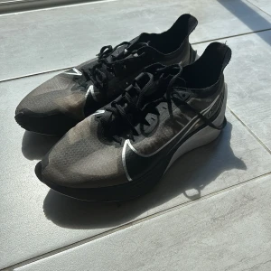 Nike ZoomX Vaporfly Next% 2 löparskor - Snygga Nike ZoomX Vaporfly Next% 2 i svart och grått med vit sula och stora vita swoosh-loggor. Skorna har en sportig, aerodynamisk design med snörning och lättviktigt syntetmaterial som andas. Perfekta för dig som vill maxa löpningen.