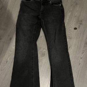 Svarta bootcut jeans Denim Day 164 - Svarta jeans från Denim Day i storlek 164. Modellen har bootcut-ben, klassiska femficksdetaljer och snygga bakfickor med lock och silverfärgade knappar. Jeansen är i ett stretchigt denimtyg och har en cool, mörk tvätt.