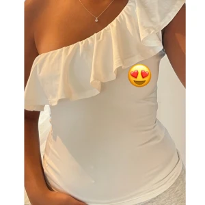 Vit oneshoulder topp med volang - Supersnygg vit topp med oneshoulder-design och stor volang längs halsringningen. Sitter tight och har en stilren, modern look som passar perfekt till jeans eller kjol. Mjuk och stretchig känsla.