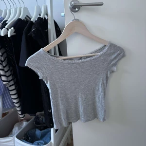 Grå croppad topp med spetskant - Superfin ljusgrå croppad topp med korta ärmar och söt spetskant runt den vida halsringningen. Perfekt för dig som gillar en enkel men ändå gullig stil. Mjuk och stretchig i bomullsmix, passar perfekt till jeans eller kjol.