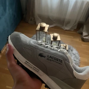 Grå sneakers från Lacoste - Snygga grå sneakers från Lacoste med vit sula och svarta detaljer undertill. Skorna har en blandning av mocka och syntetmaterial, tydlig Lacoste-logga på sidan och vita snören. Perfekta för dig som gillar stilrena och sportiga skor med coola detaljer. PASSAR BÅDE  storlek 41/42