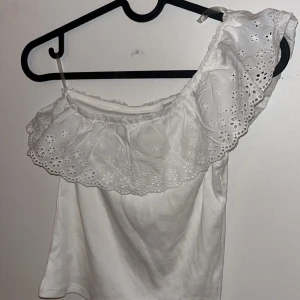 Vit offshoulder topp med volang - Superfin vit offshoulder topp med bred volangkant i broderad spets. Toppen är ärmlös, blommiga broderi. Perfekt för varma dagar och ger en söt och trendig look.