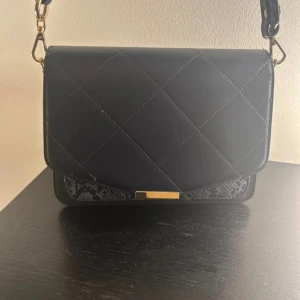 Svart quiltad axelväska från Noella - Snygg svart axelväska från Noella med quiltad design och gulddetaljer. Väskan har avtagbar axelrem i ormmönstrat material och praktiska fack inuti, inklusive ett med dragkedja. Perfekt storlek för det viktigaste och stilren look.