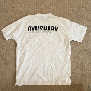Vit Gymshark t-shirt i bomull - Vit t-shirt från Gymshark med svart logga framtill och stor GYMSHARK-tryck på ryggen. Klassisk rund hals och korta ärmar. Skön och enkel modell i mjuk bomull, perfekt för träning eller chill. Snygg och stilren design som passar till det mesta.