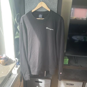 Svart långärmad tröja från Champion - Svart långärmad tröja från Champion i klassisk modell med rund hals och diskret logga på bröstet samt ärmslutet. Tillverkad i mjuk bomull, perfekt för en chill och stilren look. Passar dig som gillar enkel och sportig stil.