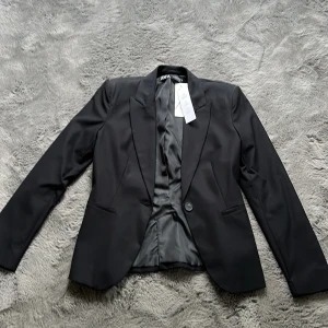 Svart kavaj - Snygg och stilren svart kavaj från Zara i storlek 34. Har lite axelvadd för en cool och strukturerad look utan att bli för stel. Klassisk modell med en knapp framtill och knappar på ärmarna funkar lika bra till vardags som när du vill klä upp dig lite. Kavajen har lappen kvar och är aldrig använd.