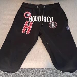 Svarta Hoodrich mjukisbyxor med patchar - Svarta mjukisbyxor från Hoodrich med flera broderade patchar och tryck i vitt och rött. Byxorna har snörning i midjan och en relaxed passform. Perfekta för en streetwear-look och chill dagar.