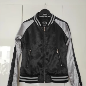 Svart och silver bomberjacka från Ecko Red - Snygg bomberjacka från EckoRed i svart och silver med glansig finish. Jackan har dragkedja framtill, två fickor med dragkedja och ribbade muddar. Klassisk collegekrage och logga på bröstet. Perfekt för dig som vill sticka ut med streetstyle! 