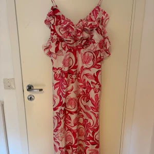 Blommig långklänning med volang - Långklänning med tunna axelband och stora volanger upptill. Klänningen har ett rosa och rött blommönster på ljus botten och är luftig och flowy. Perfekt för varma dagar när du vill sticka ut lite extra.