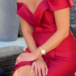 Röd axelbandslös långklänning - Snygg röd långklänning med offshoulder och v-ringad urringning. Klänningen har en figurnära passform, markerad midja med bälte och en elegant slits. Tillverkad i ett glansigt material som ger en lyxig känsla. Perfekt för bal eller festliga tillfällen.