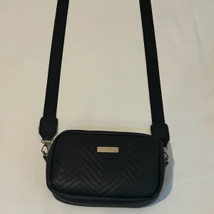 Svart quiltad axelväska med dragkedja - Snygg svart axelväska i quiltat skinnimitation med en bred rem. Väskan har ett stilrent mönster i sicksack och stängs med dragkedja. Perfekt storlek för det viktigaste och har en metallbricka framtill för en clean look.