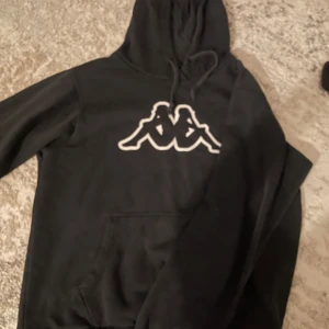 Svart Kappa hoodie med logga - Svart hoodie från Kappa med den klassiska vita loggan framtill. Tröjan har huva med snörning och en stor magficka. Perfekt för dig som gillar streetwear och vill ha en enkel men snygg look.