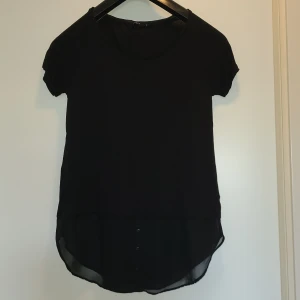 Svart blus med knappdetalj - Svart blus med rund halsringning och korta ärmar. Nederdelen är i ett tunt, transparent tyg med tre dekorativa knappar framtill. 