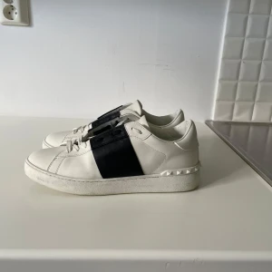 Valentino Open sneakers vit/svart - Snygga Valentino Garavani Open sneakers i vitt skinn med bred svart rem över vristen och ikoniska nitar runt hälen. Som nya i skick, Storlek 45 men passar 44. Klassisk rund tå, platt sula och vita snören. Kommer med originalkartong och dustbag. Självklart äkta!Säljer pågrund av att dem är för små och det är lågt pris för att jag vill bli av med dem. Nypris: 7000