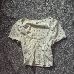 Beige blus med hålmönster från H&M - Aldrig använd på grund av att den är för stor. Supersöt beige blus från H&M med korta ärmar och knappar framtill. Toppen har ett hålmönster över hela tyget och en lätt croppad passform. Perfekt för dig som gillar romantiska detaljer och vill ha en luftig topp till sommaren.