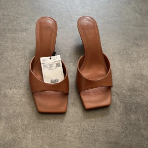 Bruna sandaler med låg klack från Mango - Snygga bruna sandaler från Mango med fyrkantig tå och låg klack. Skorna har en bred rem över foten och är tillverkade i syntetmaterial med en stilren och minimalistisk look. Perfekta för dig som gillar trendiga och enkla detaljer.