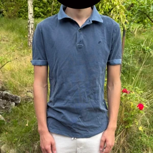 Blå pikétröja från Vaillent, stl S - Säljer en stilren blå pikétröja från Vaillent i storlek S. Tröjan har klassisk krage, korta ärmar med uppvik och knäppning framtill. Tillverkad i mjuk bomull som är skön att bära. Perfekt för en avslappnad och snygg look.