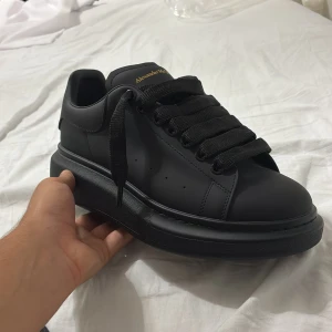 Svarta sneakers från Alexander McQueen - Stilrena helsvarta sneakers från Alexander McQueen med chunky platt sula och breda svarta skosnören. Skon är i skinn med diskret logga i guld på plösen och hälen. Snygg, minimalistisk design som sticker ut med sin kraftiga sula och exklusiva känsla. Skorna är storlek 41 men de passar även storlek 40. Helt oanvända köpte de men de var för små.
