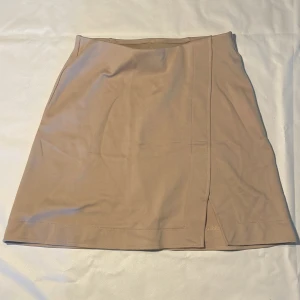 Beige kortkjol från H&M XS - Snygg beige kortkjol från H&M i storlek XS. Kjolen har en stilren omlott-look med slits framtill. Oanvänd.