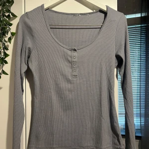 Ljusgrå/blå ribbad långärmad tröja - Ljusgrå/blå långärmad tröja med ribbad struktur och rundad, djup ringning. Tröjan har knappdetaljer framtill och är i mjuk bomullskänsla. Den har använts ett par gånger men har inga fläckar och är inte trasig någonstans😁