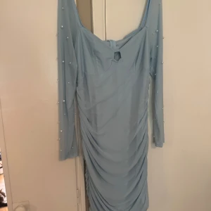 Ljusblå långärmad klänning med pärlor - Supersöt ljusblå klänning med långa ärmar och pärldetaljer. Klänningen har rynkade partier längs hela kroppen och en liten cut-out framtill. Materialet känns lätt och stretchigt, perfekt för dig som vill sticka ut lite extra.