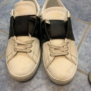 Valentino Garavani Open sneakers svart/vit - Snygga Valentino Garavani Open sneakers i vitt skinn med bred svart läderdetalj över vristen. Klassisk rund tå, vita snören och coola nitar på hälen. Perfekta för dig som gillar lyxiga och stilrena sneakers med en unik twist. STR 41!!!!!!!!!