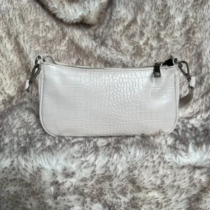 Beige axelväska i crocopräglat från Gina Tricot - Snygg beige axelväska från Gina Tricot med crocopräglat mönster och silvriga metalldetaljer. Väskan har avtagbar axelrem och stängs med dragkedja. Perfekt storlek för det viktigaste och ger en trendig touch till din outfit.