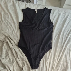 Svart ribbad omlott-body från H&M - Svart ärmlös body från H&M i ribbad bomullsmix. Snygg omlott-skärning framtill med v-ringning och tryckknappar i grenen. Perfekt att styla med jeans eller kjol för en clean look.