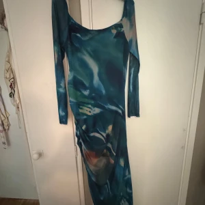 Blå långklänning med drapering - Säljer en långärmad långklänning i blått med abstrakt mönster och snygg drapering på sidan. Klänningen har en tight passform och är tillverkad i ett tunt, stretchigt material som känns bekvämt mot kroppen. Perfekt för dig som vill sticka ut med coola färgskiftningar.