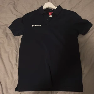 Svart pikétröja från CCM - Snygg svart pikétröja från CCM med klassisk krage och korta ärmar. Tröjan har vit CCM-logga broderad på bröstet och nacken. Perfekt för dig som gillar sportig och clean stil. Tillverkad i mjukt bomullsmaterial för skön känsla hela dagen.