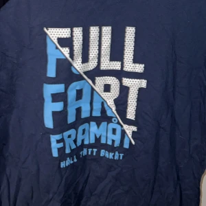 Blå DIF t-shirt med tryck 2XL - Mörkblå t-shirt från Djurgårdens IF med stort tryck på bröstet: 'FULL FART FRAMÅT HÅLL TÄTT BAKÅT' i vitt och blått. Klassisk rund hals och korta ärmar. Perfekt för dig som vill visa ditt stöd för DIF på ett stilrent sätt.