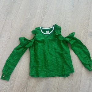 Grön blus med volang från Baum und Pferdgarten - Superfin grön blus från Baum und Pferdgarten med volangdetaljer på axlarna och öppna partier vid ärmarna. Tillverkad i en lyxig mix av siden och linne, med diskret rutigt mönster och knappar vid ärmslut. Perfekt för dig som vill sticka ut med en färgstark och trendig look.
